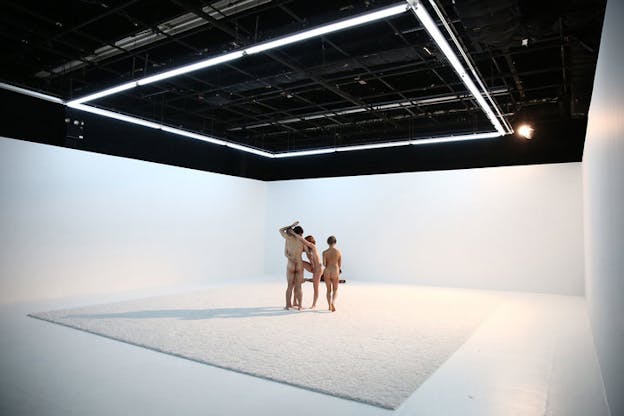 Three naked figures stand holding on to each other by tangling various limps. A fourth one stand with their back to the viewer, next to them watching two other naked figures furthest from the viewer lie on the floor.