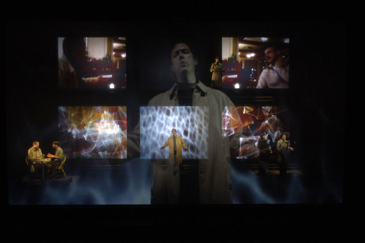 A performer stands with their arms upheld and their palms turned up in front of a black background. The image of grey smoke is behind them. Various images are overlayed on top of their image. These scenes range from images of two people talking to abstract light reflections.