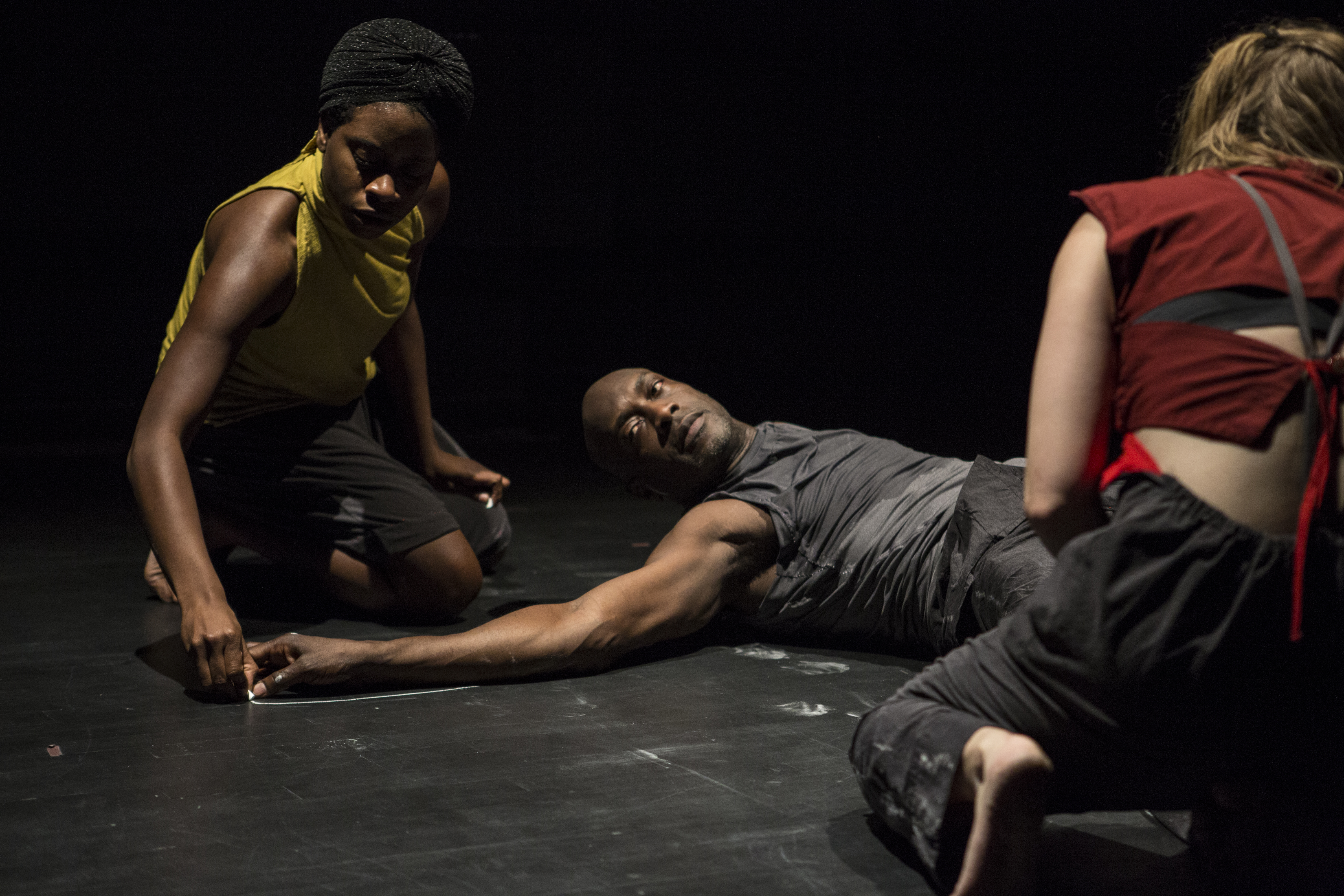 A performer dressed in all black lays on a black floor with their arm extended. Another performer dressed in yellow markes the outline of the performer's arm in chalk. A third performer dressed in red crouches in the front. 