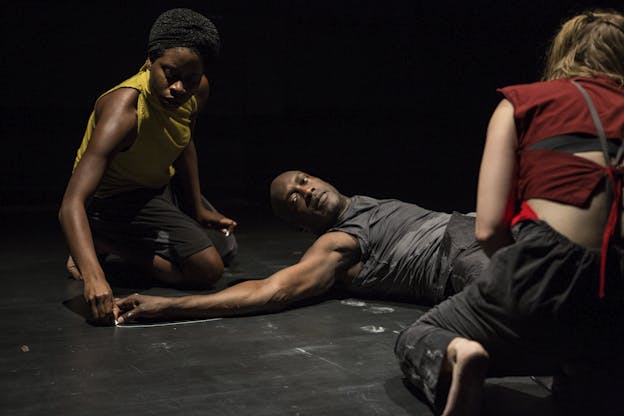 A performer dressed in all black lays on a black floor with their arm extended. Another performer dressed in yellow markes the outline of the performer's arm in chalk. A third performer dressed in red crouches in the front. 