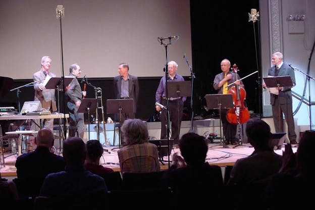 Six performers dressed in suits and button up stand on a stage behind black music stands playing their instruments. An audiences watches with their backs to the viewer.