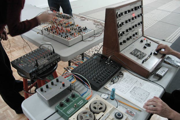 Several pieces of sound equipment are on top of grey tables. Peoples hands are visible fiddling with the equipment.