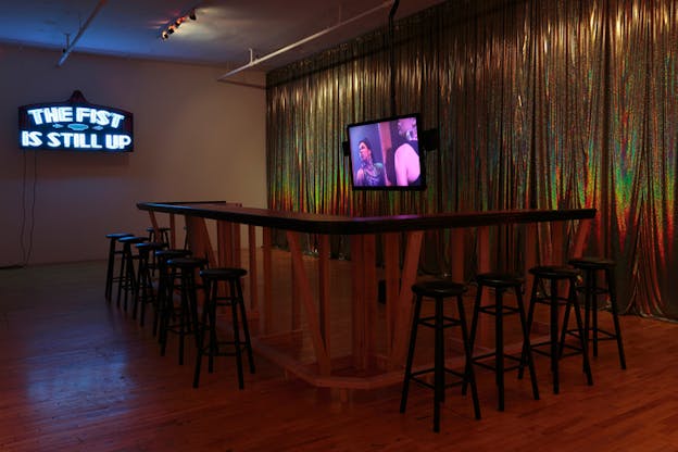 Installation view of a bar counter with empty stools and a hanging television projecting two purple-hued faces. Behind the bar is a wall of shiny gold curtains and to the left is a sign with the words lit up in blue: 