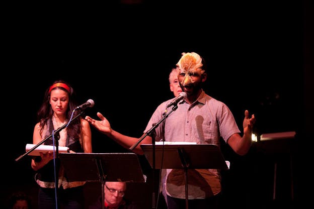 Several performers stand in a low lit space with a black backrgound. A performer in the front wears a button down shirt and a mask with a beak on it and raises their arms, speaking into a microphone. Performers in the background look down at papers or directly at the camera. 