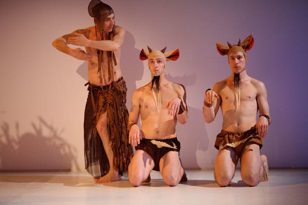 Two performers each wears a pair of goat ears, horns, and goatees headpieces and are dressed in brown puffy shorts. Their hands are raised up to mimic the forelegs of a goat while kneeling down. Another performer stands on their left and raise their bent arms laterally while looking down at the two other performers. They wear an orange-brown thigh-high slit skirt and a bonnet made of straps of lightweight fabrics. The stage has a beige floor and light background tinged with lavender color.