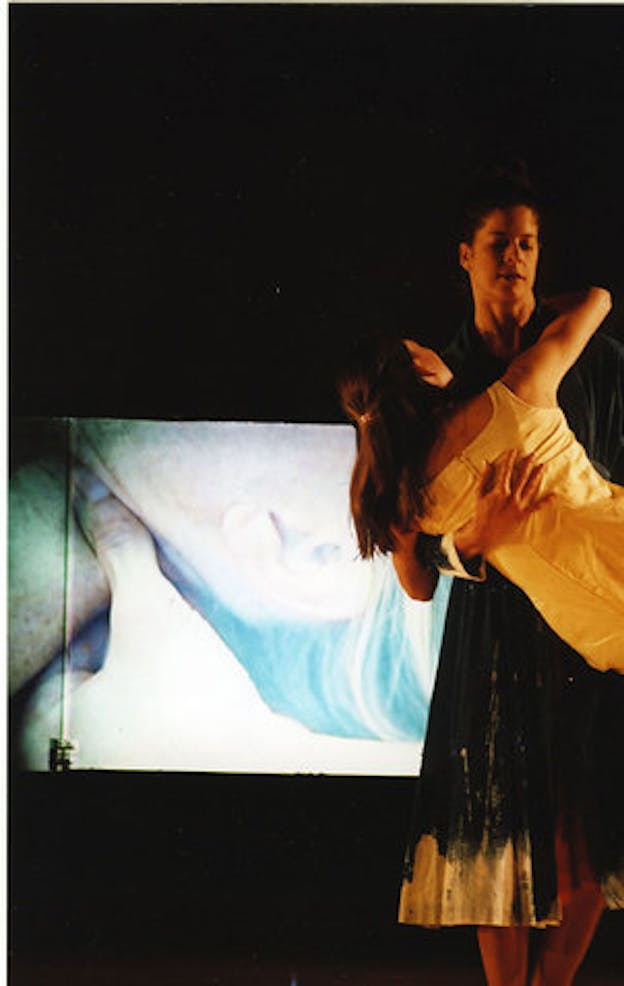 A performer wearing a blue dress holds onto a performer wearing a white dress who is leaning towards the side. The background is all black except for a blurred projection with peach and blue hues.  