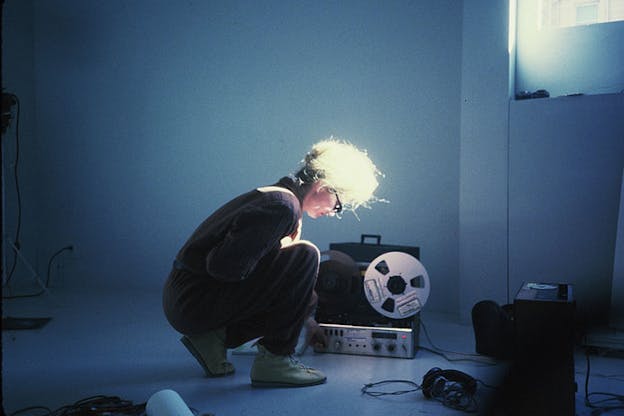 A performer crouches in a bare blue space in front of a recording machine. The performer wears their hair in a bun, has glasses, dark clothes and light shoes. The only light comes down from a window in the right wall. 