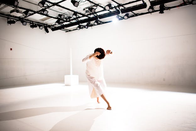 Hsiao-Jou Tang is centered, dancing in a brightly lit, bare white room. She is bent forward as she steps, head tilted down, with her elbows raised by the sides of her head. She is wearing a long sleeved, loose white dress.