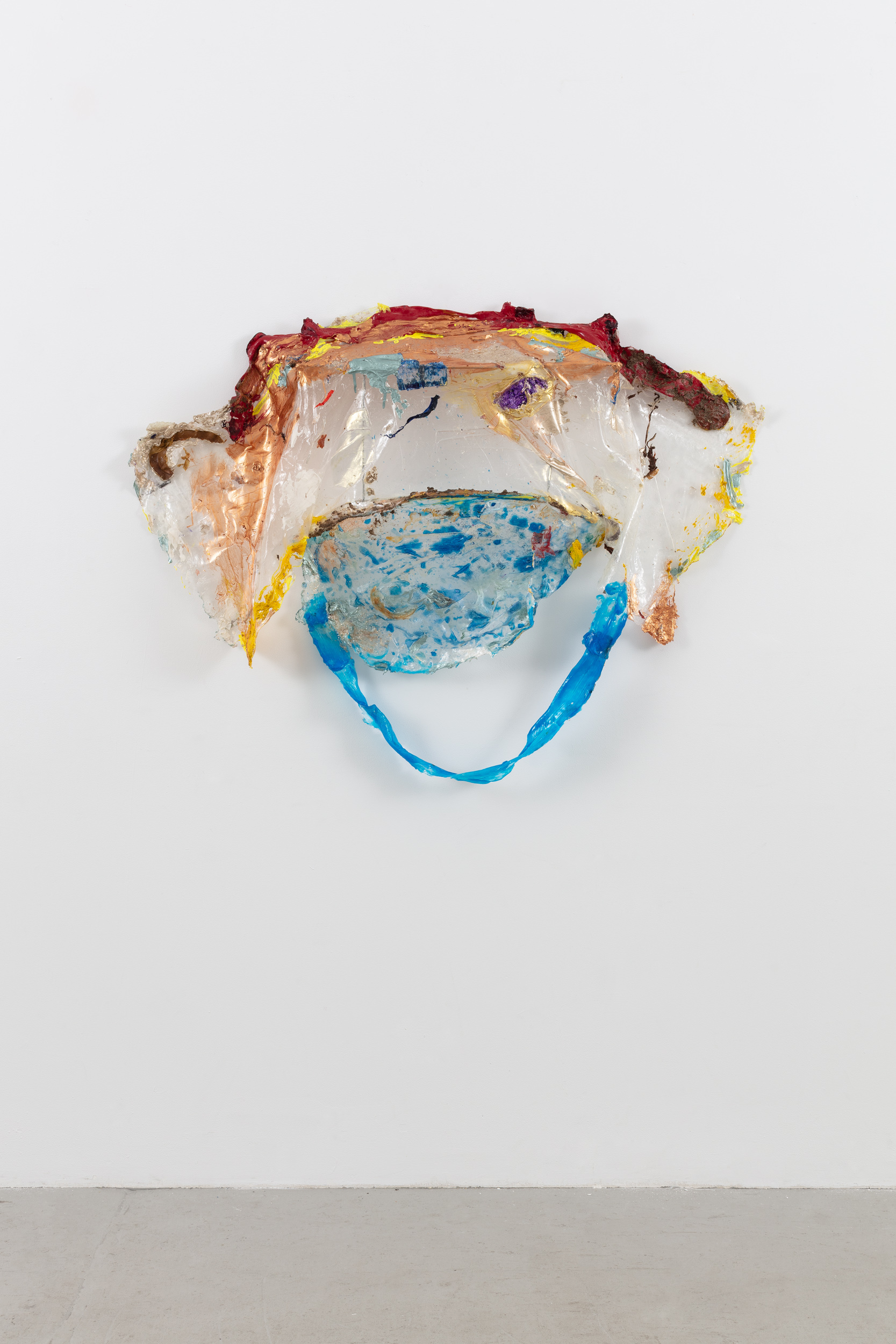 A sculpture made from torn plastic, painted with acrylic paint. The top, painted with rusty red, orange, and yellow, forms a shallow shelf covering a hanging under-section. The texture of the paint on top forms a tendril-like structure, while the under-section drapes mostly unpainted, and transitions into a smaller, round, speckled blue painted component. A strip of painted blue plastic hangs in a loose arc beneath the speckled portion.