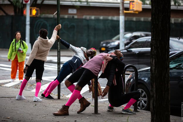 Performers with knee high neon pink socks hang by holding on to street polls and balance with their backs on bike racks near a street.