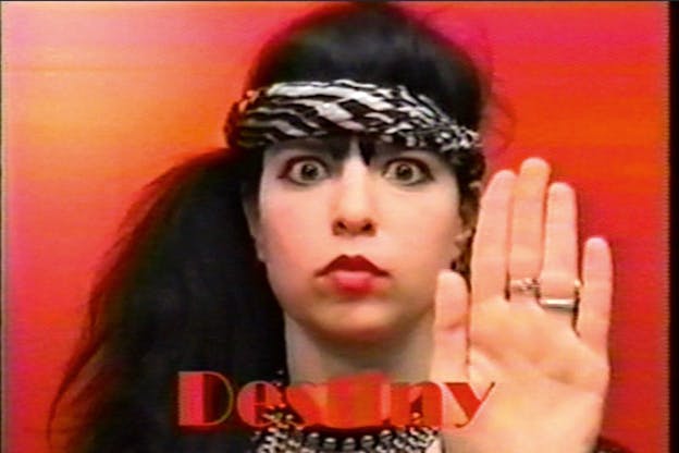 Black haired woman with red lipstick looking directly at the viewer. She wears a white and black striped bandana on her forehead and her open palm faces the viewer. The word destiny is written in red at the front. 