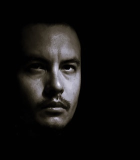 A black and white photo of aj carruthers facing the camera with his gaze looking upwards. Only his face is visible in an otherwise dark space, brows slightly furrowed.  