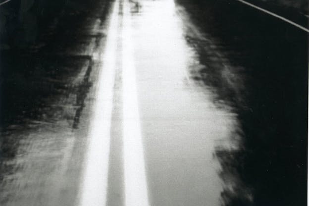 Black and white photgraph centering on double white road markings. Black blurry forms present themselves at the left and right of the road.