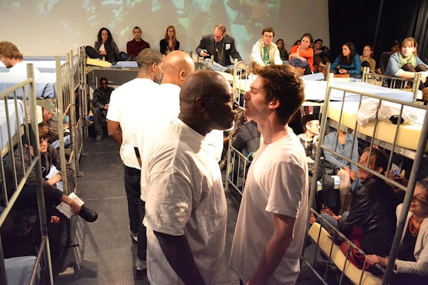 Multiple people sit on bunker bed surrounding performers dressed in white shirts in the middle. Two of the performers closest to the viewer stand sideways looking at each other.