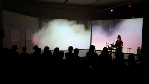 A photograph of Pamela Z performing, standing in front of a music stand and microphone. Behind her, a screen projection depicts light green and light pink smoke. On the far left of the screen, there are two windows on a light grey facade pictured. Pamela Z is illuminated by pink light but the rest of the scene is dark and the audience who sits in front of her, watching, is enclosed in shadows. 