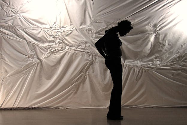 Okpokwasili standing sideways, bending their neck forward and holding their arms behind their hips, silhouetted against a wall of white bunched sheets.