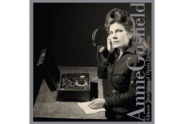 A black and white image of Gosfield sitting at a table with a box open in front of her with many knobs and wires attached to it. She holds a pair of headphones up to her ear and looks directly at the camera. With her other hand, she writes something on the piece of paper on the table. She wears her hair in an elaborate up-do and wears a dark colored jacket with many pockets. On the right, the words 