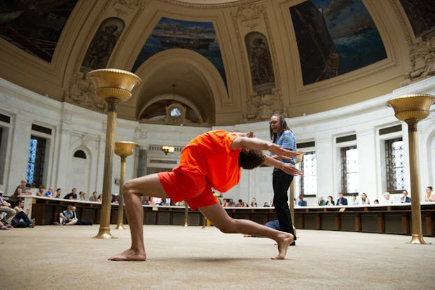 A figure clad in orange sideways to the viewer leans their body back while kneeling, their arms above their head extend back. Behind them a standing person talks and in the background people sit behind a semi-circular table surrounding them.