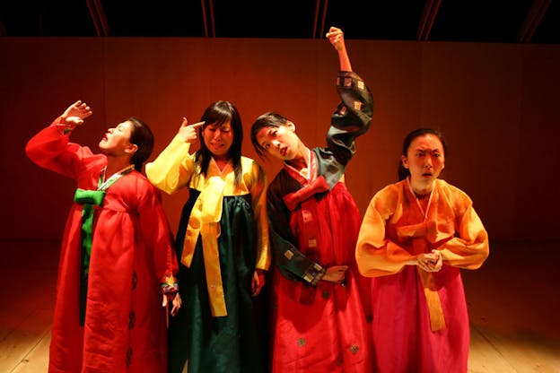 Four people dressed in traditional Korean wear are each posed differently: From the left, the first person mimes drinking a glass of liquid. The second one mimes holding a gun to their head. The third one mimes hanging themselves. The last one mimes stabbing themselves. The set has wood floors and walls.