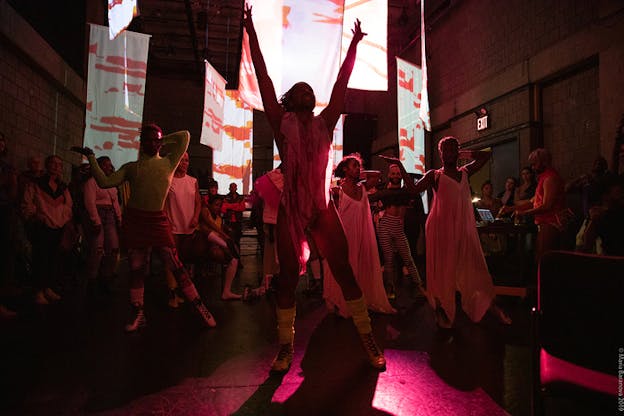 Performers dressed in white stand in formation in a red lighted room with one hand to their head  and the other extended to the side, as the audience behind watches. The performer in the front closest to the viewers has their hands extended upwards. White banners hanging from the ceiling stand behind and on top of them, a projection of white and orange lines and circles on them. 