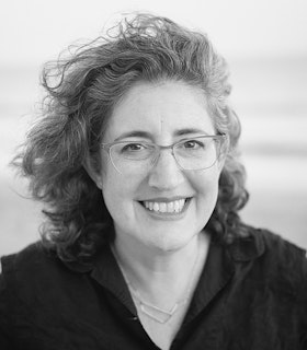 A black and white portrait of Marcella Durand with shoulder-length wavy hair tousled to one side, wearing thin-framed glasses and a black collared shirt.