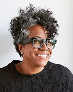 Portrait of a smiling Jennie Jones with short dark curly hair with a gray streak on the right front side, big square black glasses, and a textured black shirt.