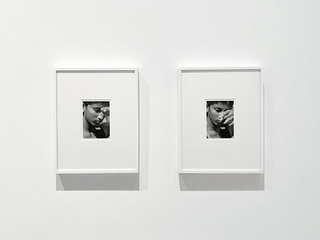 A photograph of two black and white framed photographs in white frames hung next to each other. Surrounded by wide white mat within the frame, the photographs are closeup shots of a subject's face. In both, the subject gazes downwards, shielding their eyes from the sun.