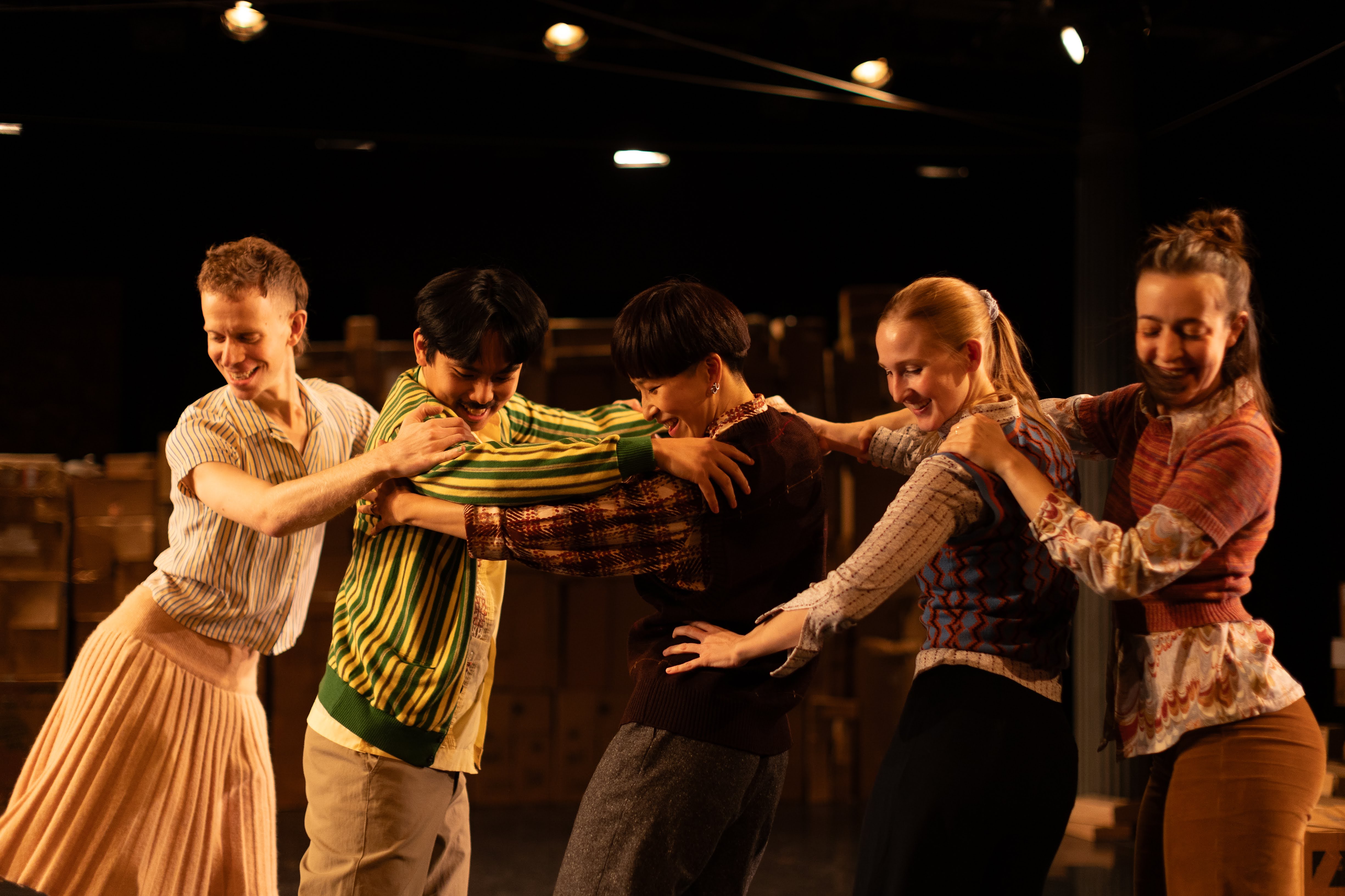 Five performers dance in a line in a warmly-lit space, embracing each other and smiling as they gaze at the floor, all wearing patterned, colorful clothing. Hsiao-Jou is centered in the frame, she stands with her body facing the left, arms raised and holding the torso of another dancer facing her. Two dancers stand behind her on the right half of the frame, facing the same direction with their hands resting on the next's shoulders. Directly across from Tang on the left side of the frame are two dancers, both facing Tang with their hands on the person in front's shoulders or hips. 