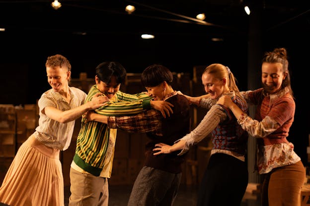 Five performers dance in a line in a warmly-lit space, embracing each other and smiling as they gaze at the floor, all wearing patterned, colorful clothing. Hsiao-Jou is centered in the frame, she stands with her body facing the left, arms raised and holding the torso of another dancer facing her. Two dancers stand behind her on the right half of the frame, facing the same direction with their hands resting on the next's shoulders. Directly across from Tang on the left side of the frame are two dancers, both facing Tang with their hands on the person in front's shoulders or hips. 