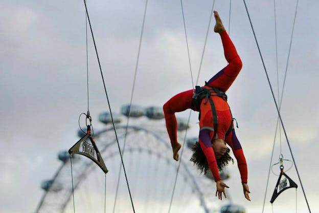 A performer clad in red hangs upside down in the air by a supportive system surrounding their waist. Behind them the top of a ferris wheel.
