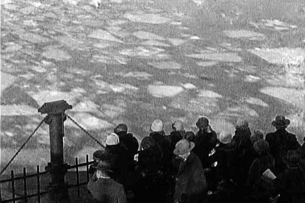 A black and white video still of people huddled together behind a fence looking down at water with ice chunks floating in it.