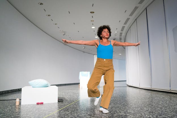 Performer in a blue tank top and sand brown pants lifts one leg up while keeping both arms raised to the sides. Behind them a white stand with a blue pillow and pink glasses at its bottom.