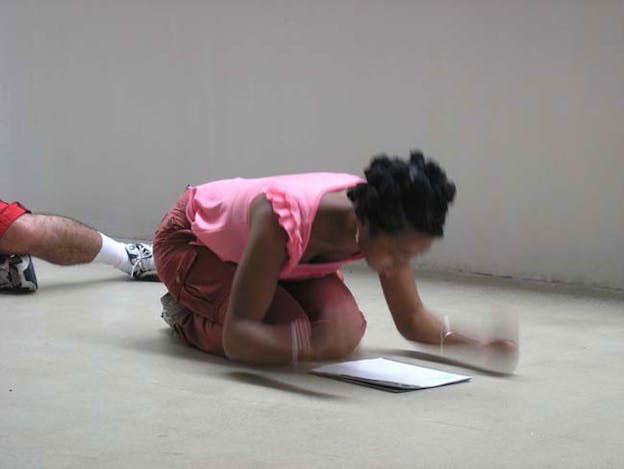 One person kneels down on a white floor and pounds the floor with their hands while looking at a stack of paper. 