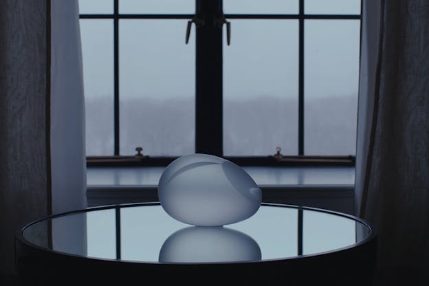 A close up of white spherical structure with indentations in forms of a slice and wave, on a dark glass surface in front of a window.