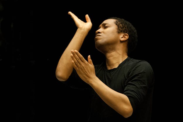 Harrell in a black tee-shirt set against a pitch black backdrop closes their eyes expressively, lifts their forearms, and curves their hands.