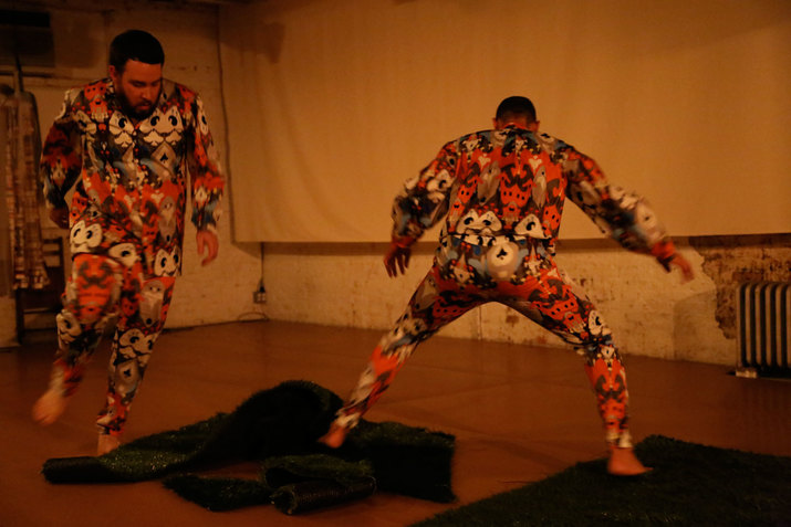 Two performers dressed in colorful pajamas move in a yellow warmly-lit room, their bodies slightly blurred in motion.