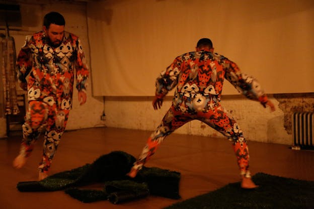 Two performers dressed in colorful pajamas move in a yellow warmly-lit room, their bodies slightly blurred in motion.