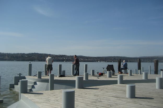 People sit and support themselves on gray cylinders placed though-out a wooden wharf.