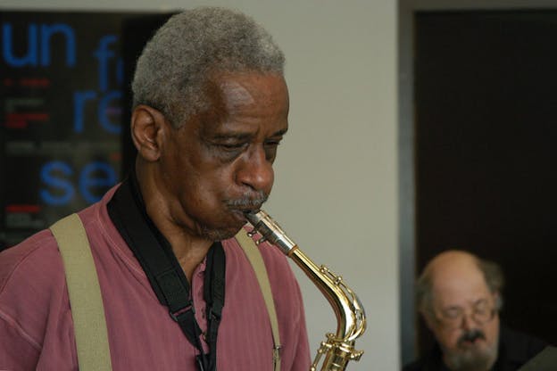 Photograph of a person stopping at their shoulders. They can be seen with puffed cheeks playing a brass instrument. 