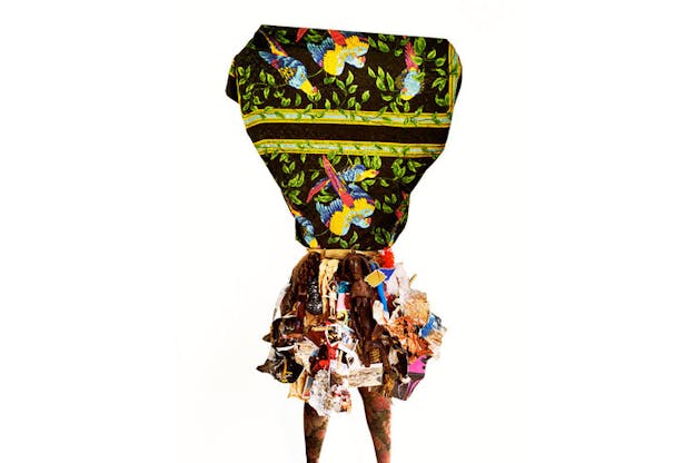 Collage sculpture of mixed materials including parrot-decorated fabric, floral tights, wooden statues, feathers, and crumpled glossy paper against a white backdrop.
