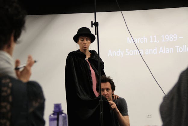 Person in a black bowler hat and black cape rests their hand on the shoulder of a person whose head is at their waist. In the back projected white text against a taupe wall reads: 