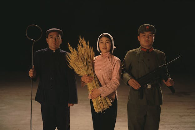 A performance still of three performers looking directly ahead in an otherwise bare spare. The performer on the left wears a blue jacket, blue pants, and carries a thin metal rod twisted into a circle at the top. The performer in the middle wears a white bonnet, red gingham shirt and holds a bundle of wheat. The performer all the way to the right wears a military uniform and holds a gun. 