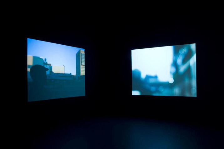 Two dusky blue hued images, one depicting the silhouette of the back of a person's head in an urban setting and one depicting out-of-focus buildings, suspended in pitch-black space. 