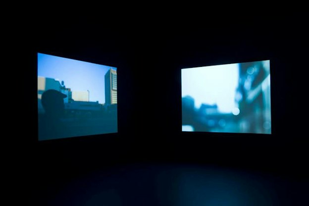 Two dusky blue hued images, one depicting the silhouette of the back of a person's head in an urban setting and one depicting out-of-focus buildings, suspended in pitch-black space. 