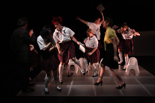 Seven performers wearing white button-down tops with plaid skirts are in various stages of skipping, stomping, pacing, and waving their hands in the air. They are standing on a gridded platform with small white plastic chairs surrounding them.