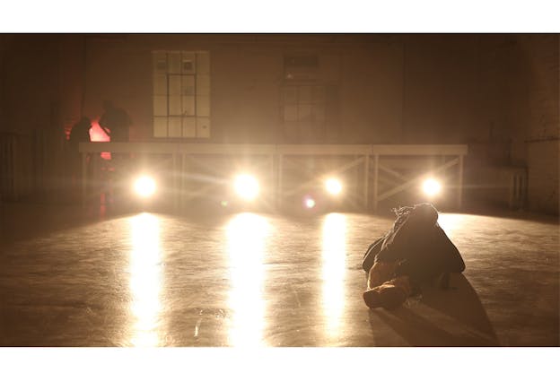 A figure lies down on a stage floor with the bottom of their  feet facing the viewer. Behind them four strong lights create lines of the stage.