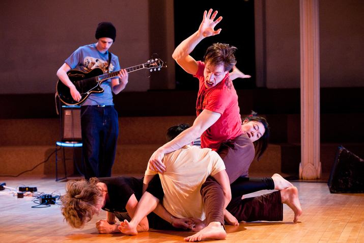 Performers tangle on the floor as a musician behinds them plays the electric guitar. 