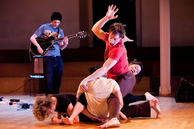 Performers tangle on the floor as a musician behinds them plays the electric guitar. 