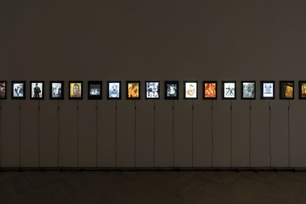 Aligned lightboxes depicting various images situated on a wall and connected each by cables going straight to the floor. They consist of faces and figures depicted in cold and warm tones.
