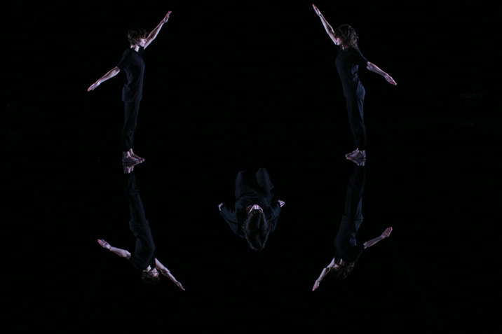 Two performers dressed in black stand symmetrically facing each other and raising one arm in front and one in back with one performer lying with their back on the floor between them in a black room with a mirrored floor reflecting their bodies. 